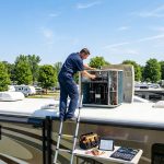 Mobile RV technician servicing a rooftop air conditioner on a fifth wheel at a campground during hot summer weather.